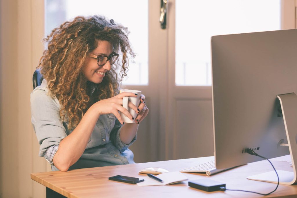 לעבוד בתחום QA - אישה מחייכת מחזיקה כוס קפה ועובדת על המחשב 